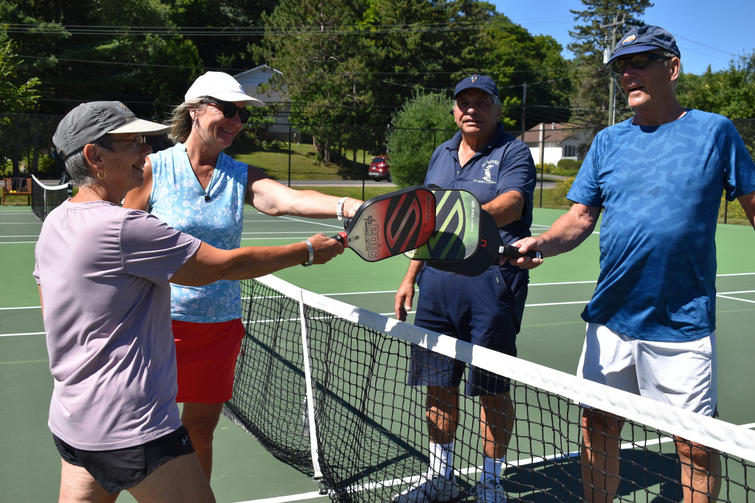 Four older adults playing pickleball on an outdoor court in Huntsville, two on each side of the net, with their paddles touching in the center.